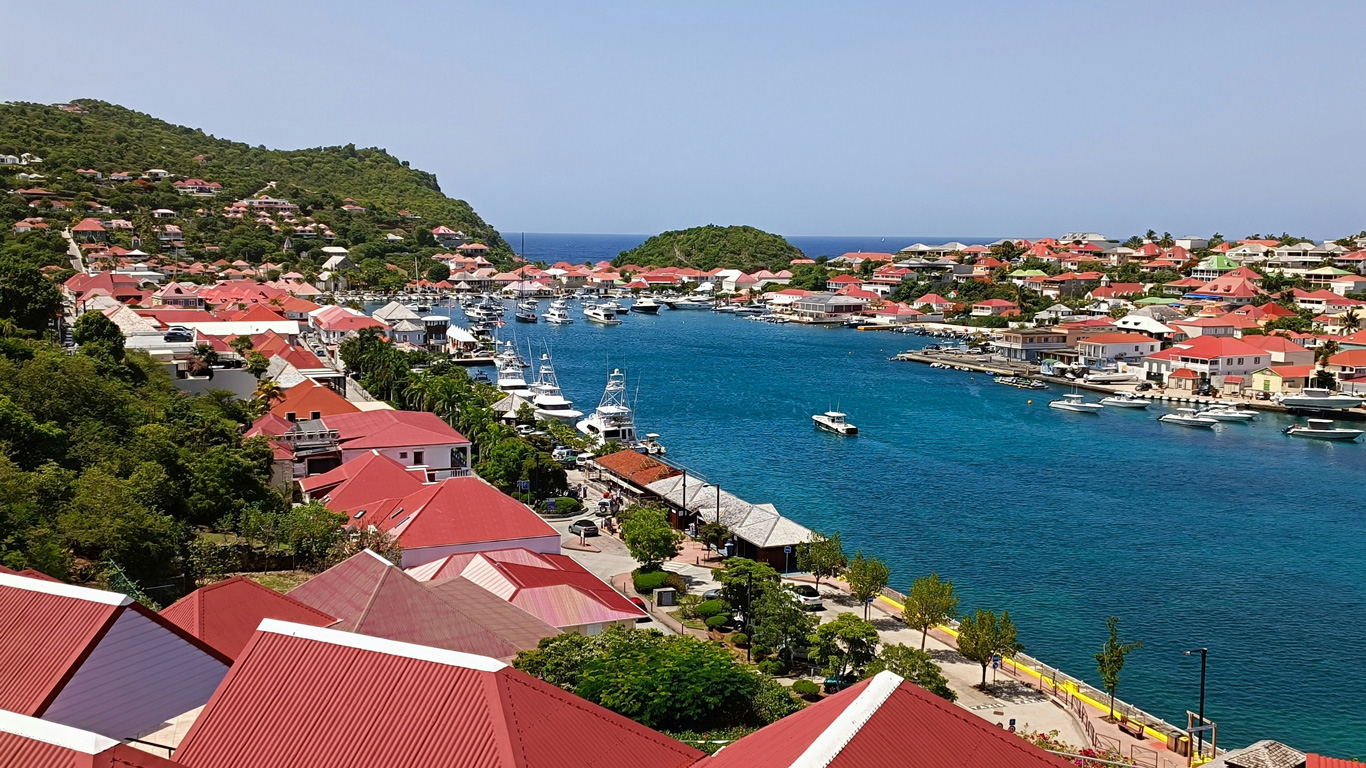 Una vista panorámica de Gustavia, la capital de Saint-Barthélemy, con sus característicos techos rojos y casas coloridas rodeando un puerto de aguas turquesas. Numerosos yates y barcos están atracados a lo largo del muelle, reflejando el ambiente lujoso y exclusivo de la isla. Las colinas verdes en el fondo contrastan con la arquitectura costera, creando una escena vibrante y pintoresca del Caribe francés.