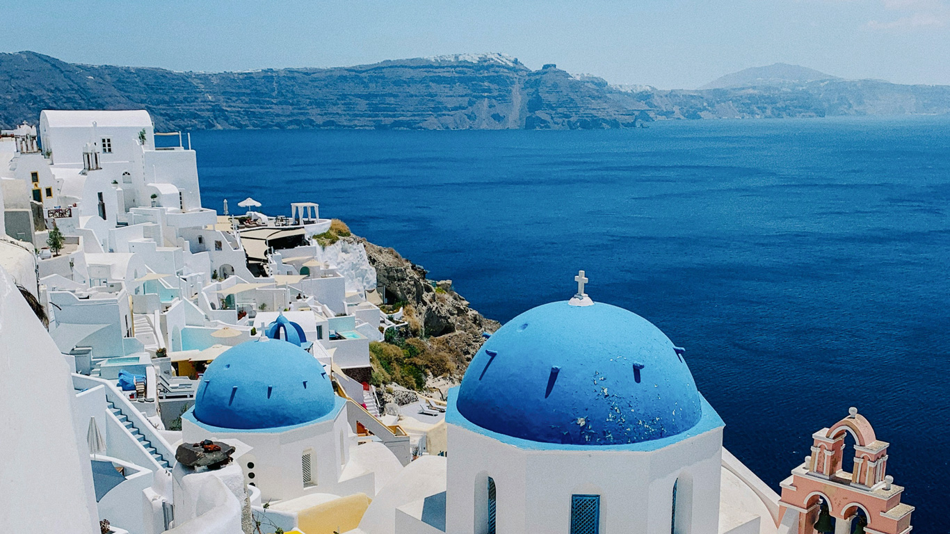 Una vista icónica de Santorini, Grecia, con sus distintivas iglesias de cúpulas azules contrastando con las fachadas blancas de las casas en la ladera. Al fondo, el mar Egeo de un azul profundo se extiende hasta el horizonte, con acantilados volcánicos en la distancia. La arquitectura tradicional y el paisaje espectacular crean una escena pintoresca y emblemática de las islas griegas.