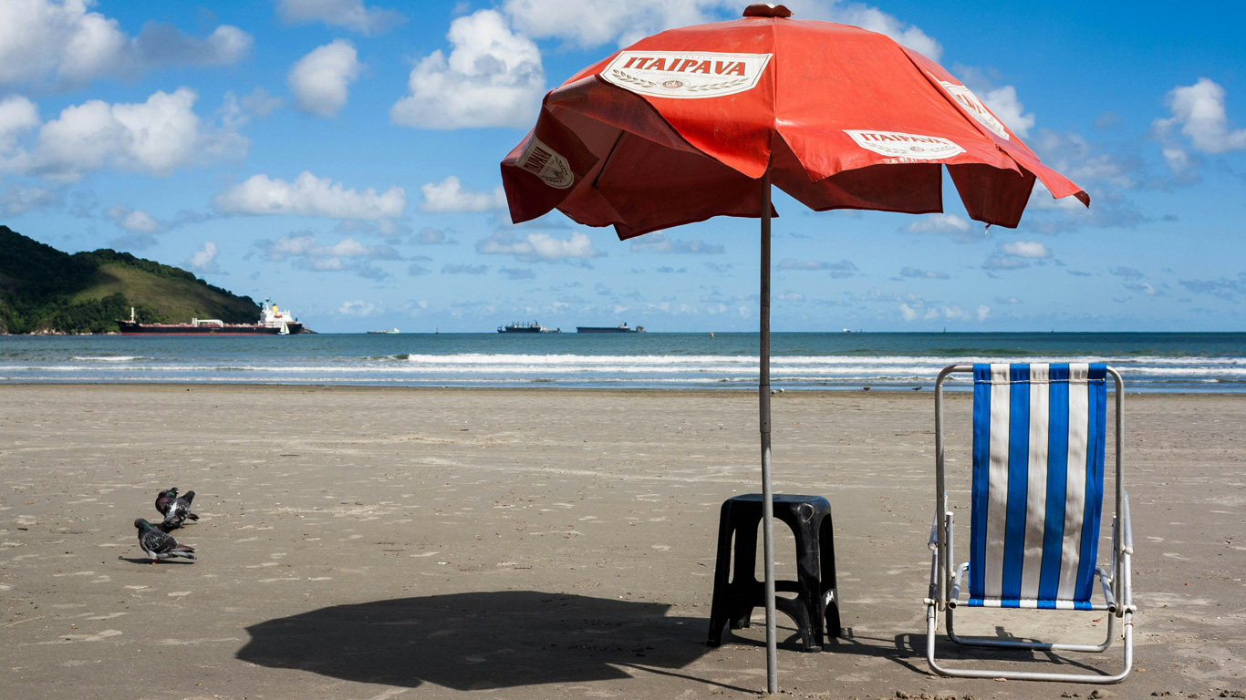 Una sombrilla roja con el logo de Itaipava brinda sombra a una silla de playa de rayas azules y blancas en la arena de una playa en Santos, São Paulo. En el fondo, el mar azul se extiende hacia el horizonte con varios barcos y un paisaje montañoso. La escena transmite una sensación de relajación y tranquilidad en un día soleado junto al océano.