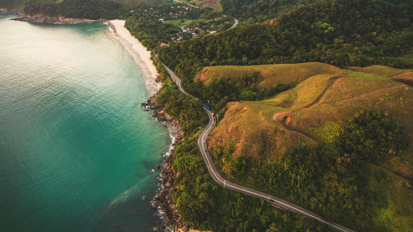 Vista aérea de una carretera sinuosa que bordea la costa, rodeada de colinas verdes y un mar turquesa en São Sebastião, São Paulo. La playa de arena blanca contrasta con la densa vegetación y las pequeñas casas visibles en la distancia. La imagen captura la belleza natural y la tranquilidad de este destino tropical.