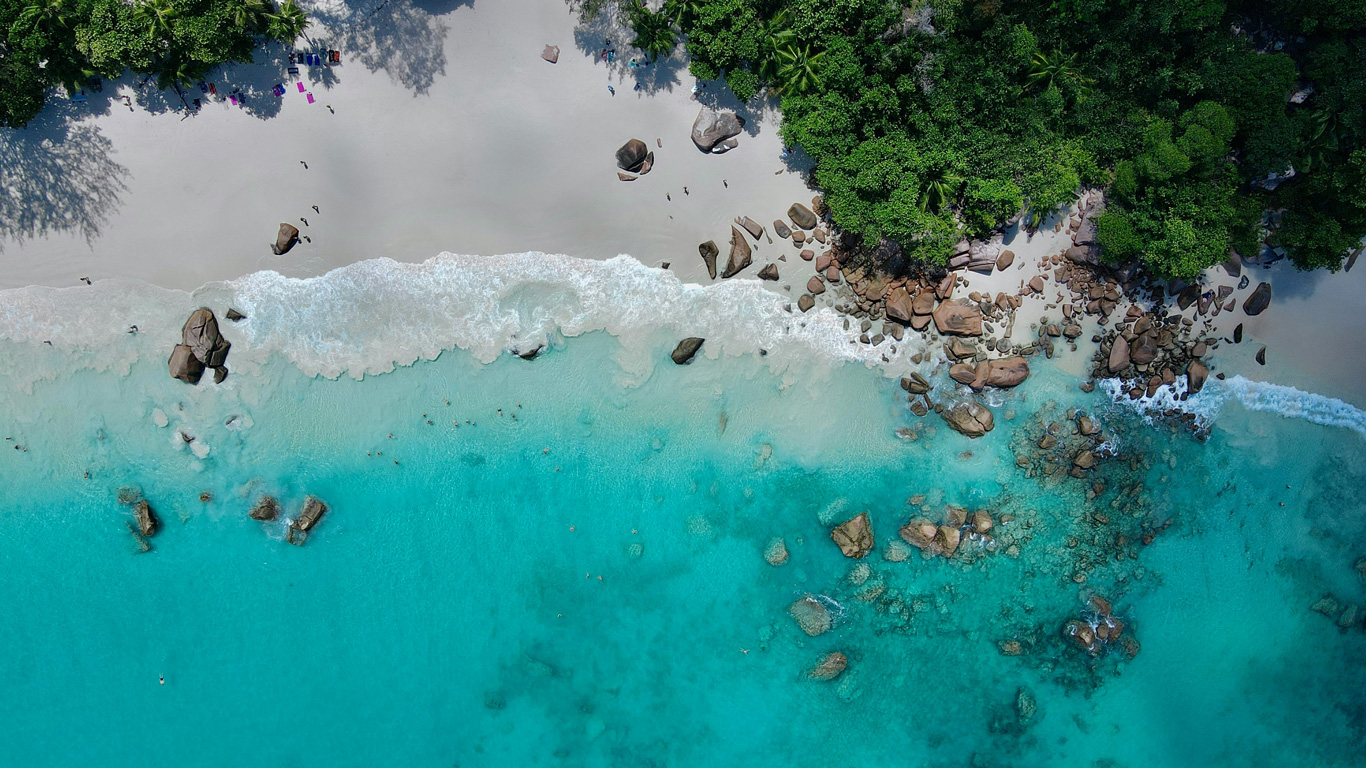 Una vista aérea de una playa paradisíaca en Seychelles, con arena blanca inmaculada, aguas cristalinas de color turquesa y grandes formaciones rocosas a lo largo de la orilla. Las olas suaves crean una espuma blanca mientras bañistas disfrutan del mar, y una densa vegetación tropical enmarca el paisaje. La combinación de colores vibrantes y la naturaleza prístina hacen de este un destino verdaderamente idílico.