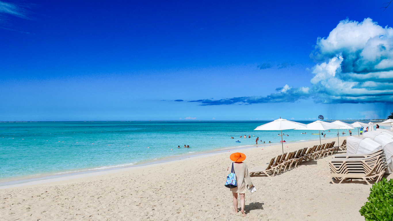 Una playa paradisíaca en las Islas Turcas y Caicos, con arena blanca y aguas cristalinas en tonos turquesa. En primer plano, una mujer con un sombrero naranja camina descalza hacia el mar, mientras varias tumbonas y sombrillas blancas están alineadas en la orilla. El cielo azul intenso con nubes esponjosas añade un toque espectacular a esta escena tropical de ensueño.