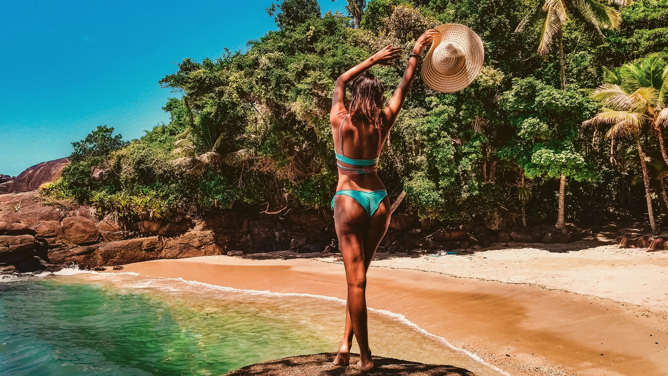 Una mujer con bikini azul turquesa se encuentra de espaldas sobre una roca, levantando un sombrero de paja en una mano mientras disfruta de la vista de una playa paradisíaca. El agua cristalina y la arena dorada contrastan con la exuberante vegetación y las palmeras al fondo. La escena transmite una sensación de libertad, aventura y conexión con la naturaleza en un destino tropical.