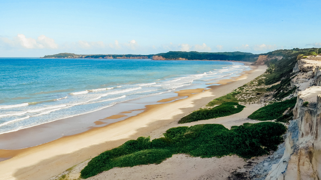 Playa-de-Cacimbinhas Vista panorámica de la Playa de Cacimbinhas, con acantilados blancos y vegetación verde contrastando con la arena dorada y el mar azul. La playa se extiende a lo largo de la costa en un paisaje natural y casi desierto.