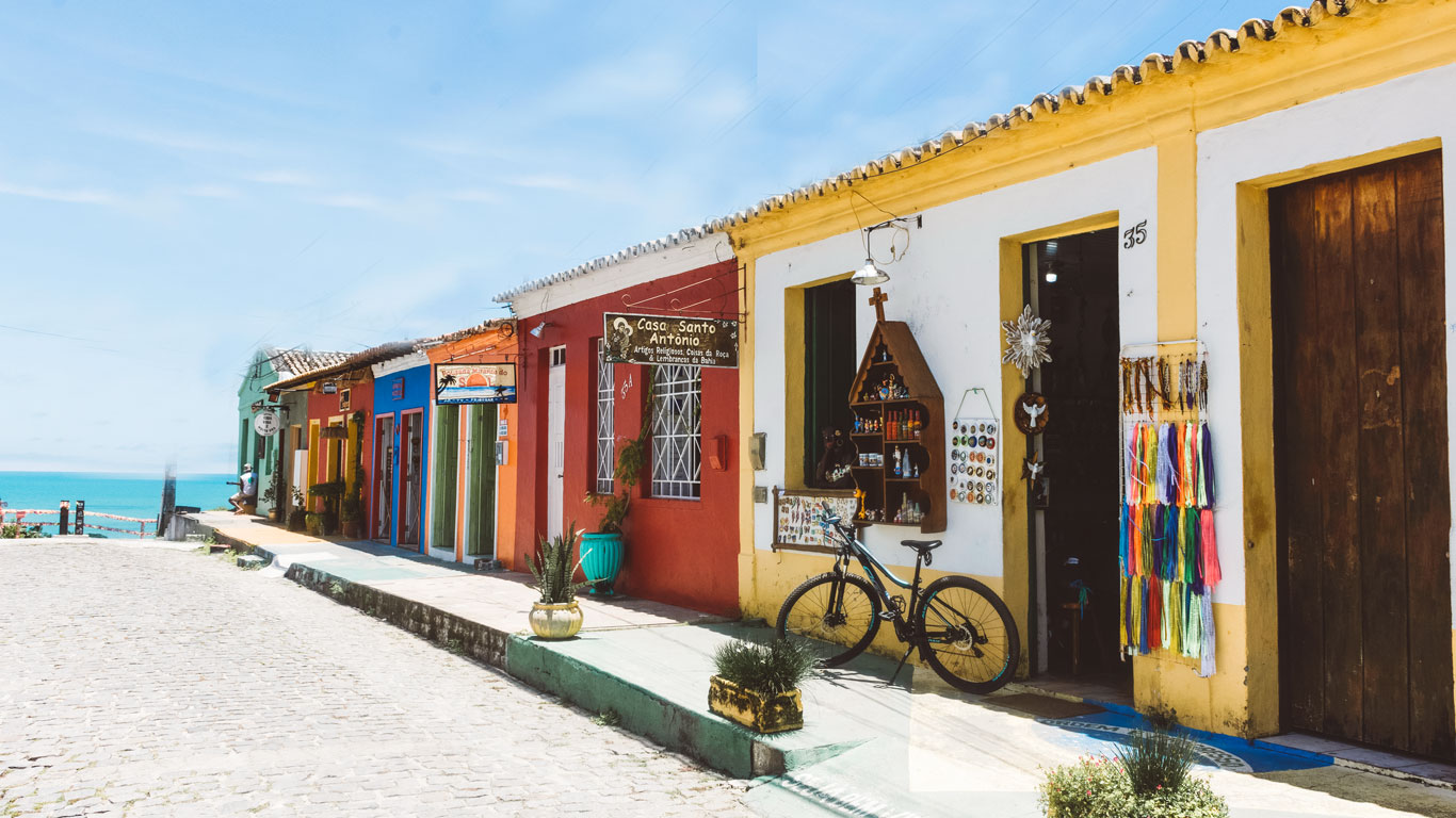 donde-alojarse-en-arraial-dajuda-bahia Calle empedrada junto al mar con casas bajas de colores vivos como rojo, amarillo, verde y azul, decoradas con plantas, carteles de artesanía y una bicicleta apoyada en la acera. El cielo está despejado y se ve el océano al fondo.