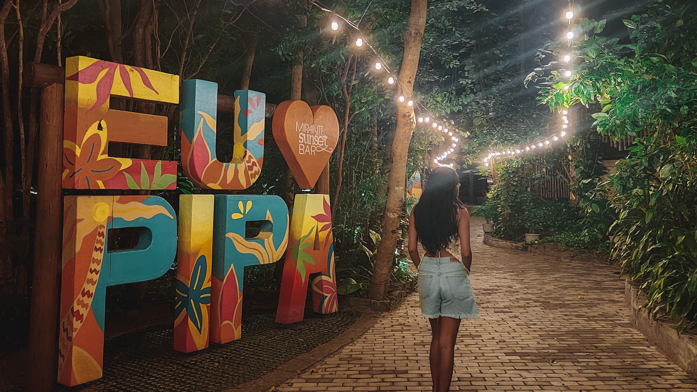 noche-en-pipa Escena nocturna con una mujer caminando por un sendero adoquinado iluminado por guirnaldas de luces, al lado del colorido cartel “Eu amo Pipa” junto a árboles frondosos. Ambiente acogedor y lleno de encanto tropical en el corazón de Pipa.