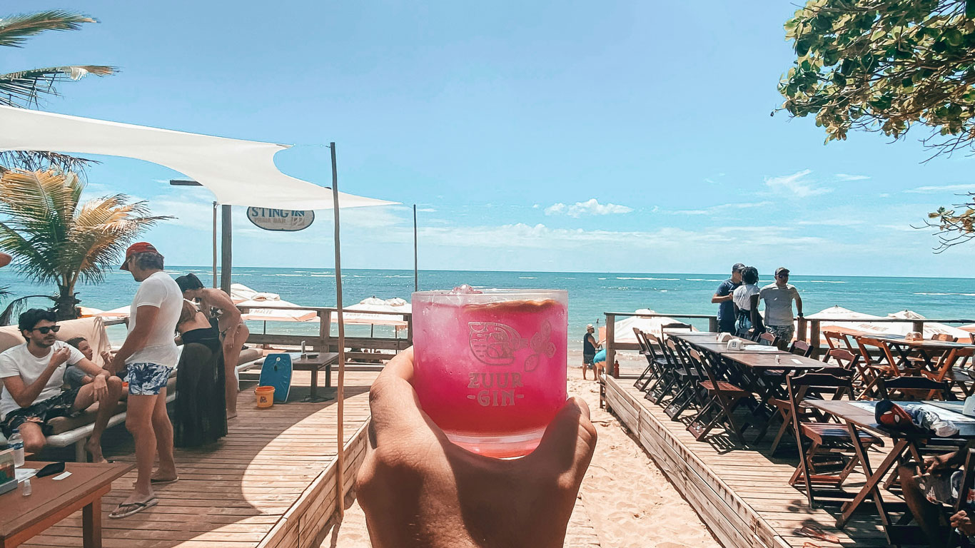 playa-de-aracaípe-arraial-ajuda Ambiente animado en un chiringuito de playa con vistas al mar, donde personas disfrutan de bebidas refrescantes y cócteles como un gin rosa, bajo sombrillas blancas y toldos de lona. Perfecto para quienes desean saber dónde alojarse en Arraial d'Ajuda, Bahía y disfrutar de un ambiente relajado junto al mar.