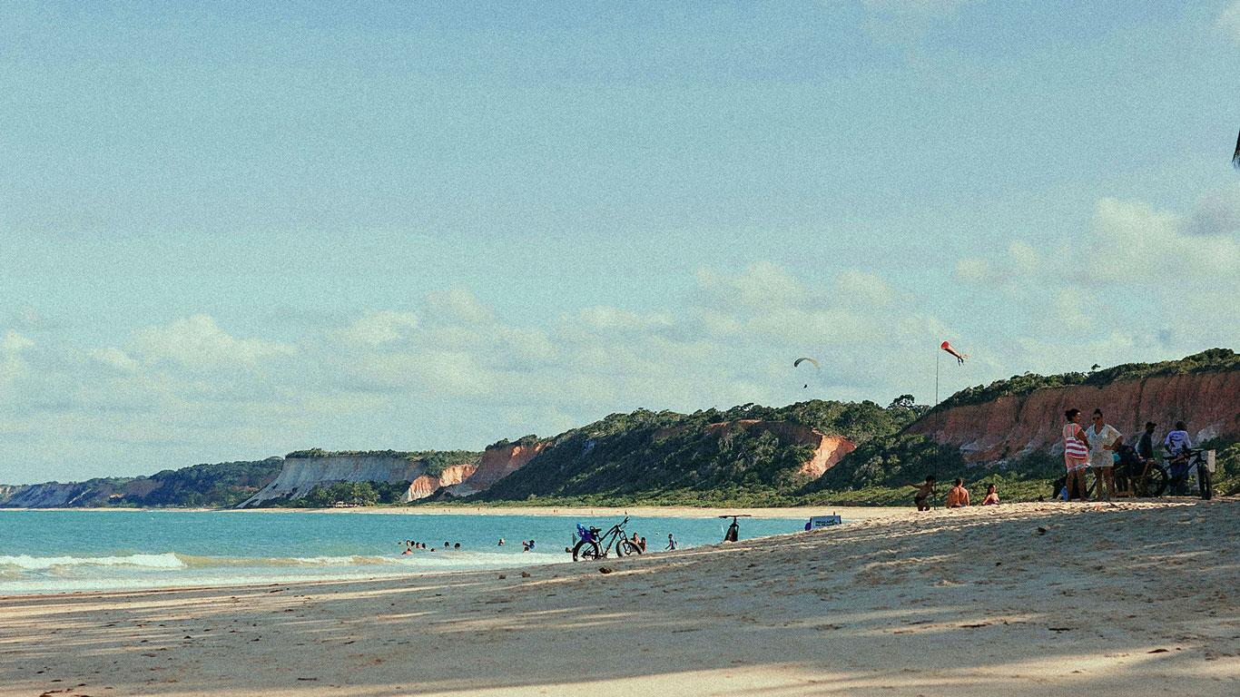 playa-de-pitinga-arraial-ajuda Vista panorámica de una playa amplia con arena clara, bordeada por altos acantilados rojizos y vegetación exuberante, donde varias personas disfrutan del mar y se observan parapentes en el cielo. Esta playa es una excelente opción para quienes buscan dónde alojarse en Arraial d'Ajuda, Bahía, rodeados de naturaleza y aventura.