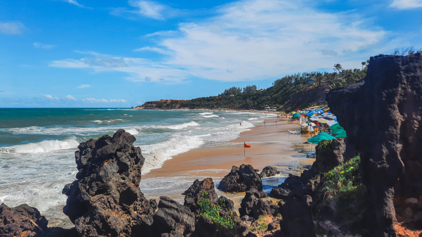 playa-do-amor-pipa-2 Vista panorámica de Playa do Amor en Pipa, Brasil, con olas rompiendo sobre la orilla, formaciones rocosas oscuras en primer plano y sombrillas de colores en la playa. Al fondo se ven acantilados y una línea de árboles bajo un cielo azul claro con algunas nubes.