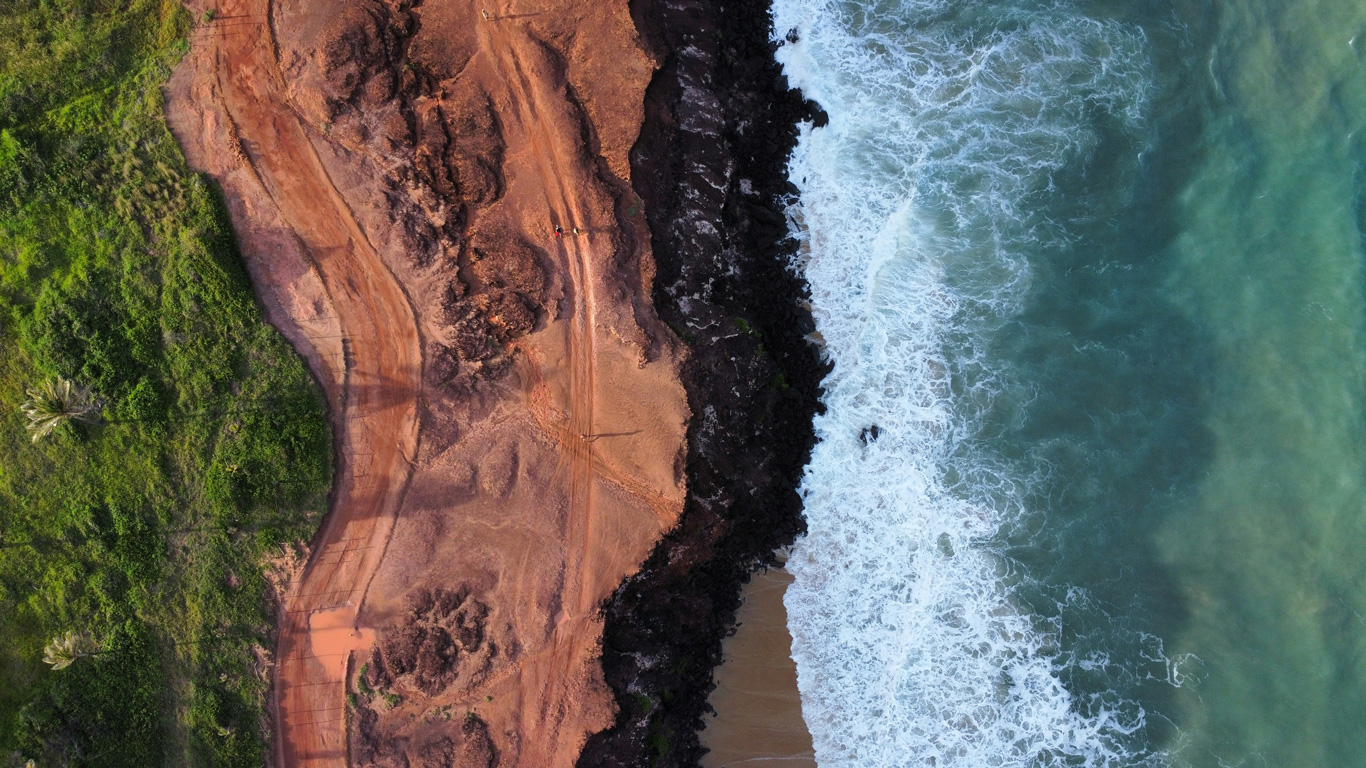 praia-de-pipa Imagen aérea de la costa de Pipa que muestra un contraste visual entre vegetación verde, terreno rojizo y la espuma blanca del mar golpeando las rocas negras. Un sendero de tierra recorre el borde del acantilado hasta la orilla.