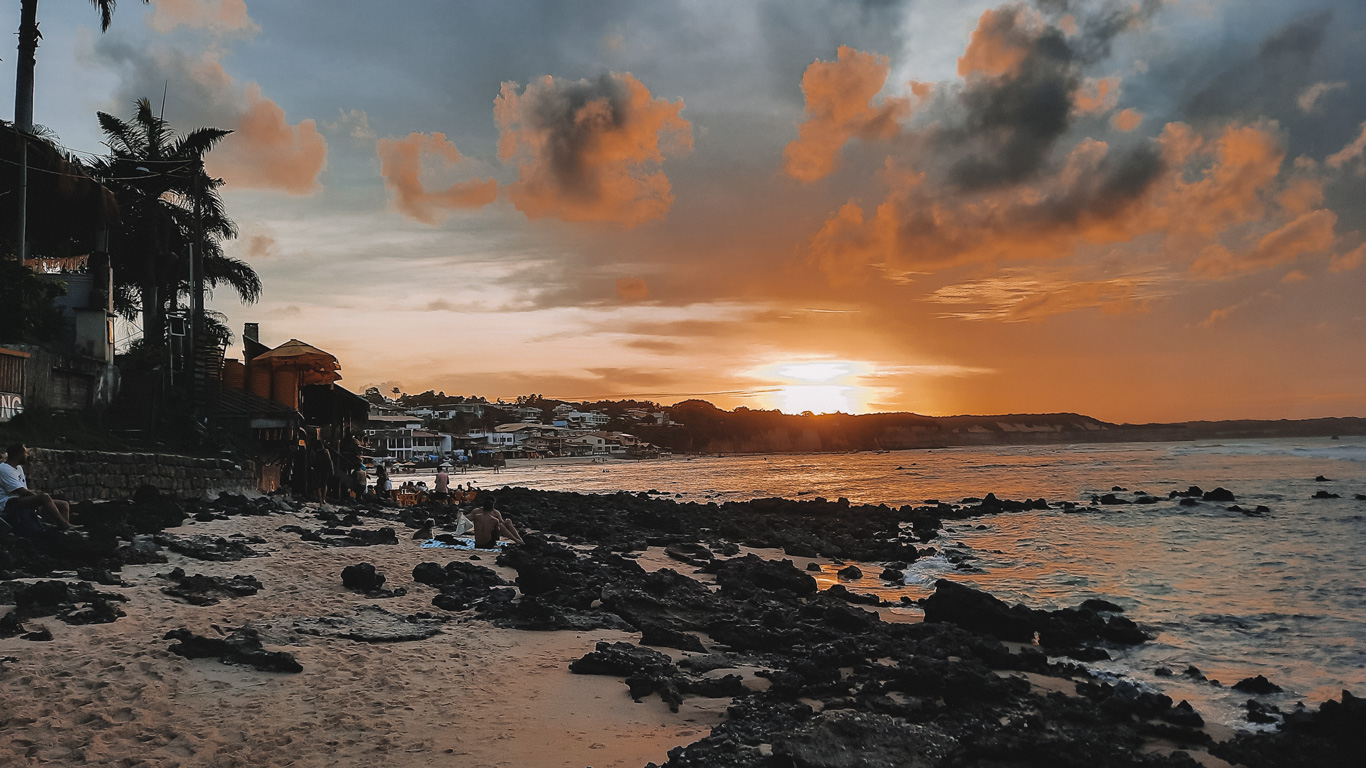 puesta-del-sol-en-pipa-1 Atardecer dorado en la playa de Pipa con cielo parcialmente nublado y tonos anaranjados reflejados en el mar. Varias personas se sientan entre las rocas oscuras observando la puesta del sol frente al horizonte.