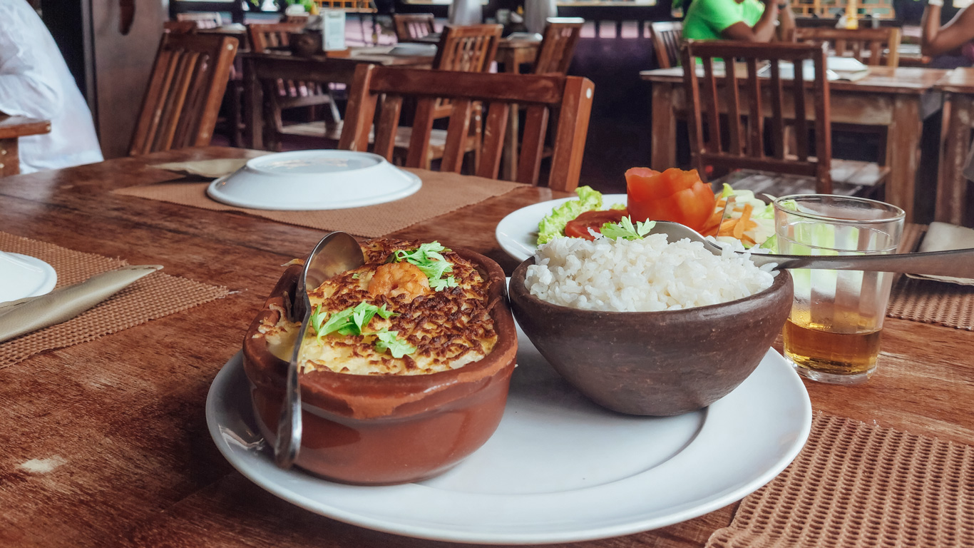 restaurantes-en-pipa Plato típico brasileño servido en un restaurante rústico con mesas de madera, compuesto por arroz blanco, una especie de gratinado de carne o pescado cubierto de patatas ralladas, ensalada fresca y una bebida en vaso. El ambiente es acogedor y relajado, ideal para disfrutar de la gastronomía local.