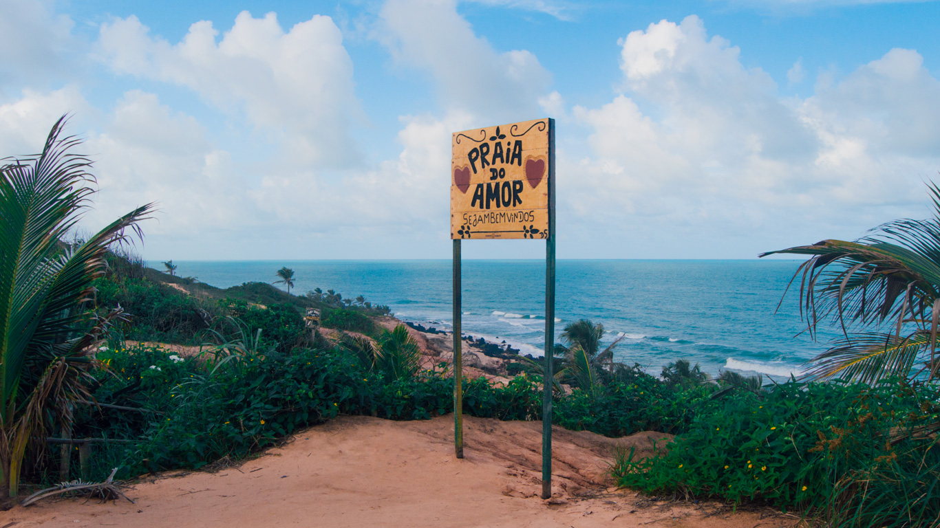 tour-en-buggy-pipa Cartel artesanal de madera en lo alto de un acantilado anunciando “Praia do Amor, Sejam Bem-vindos”, con corazones decorativos. Al fondo, el mar azul y el cielo nublado enmarcan el paisaje lleno de vegetación tropical.