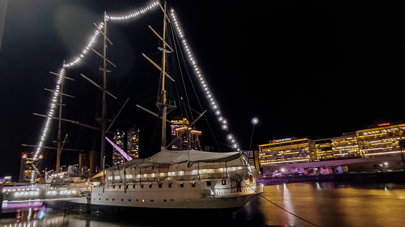 Fragata-Sarmiento El barco Fragata Sarmiento anclado en Puerto Madero, iluminado con luces a lo largo de los mástiles, reflejándose en las aguas tranquilas. Edificios comerciales brillan al fondo en contraste con la noche.