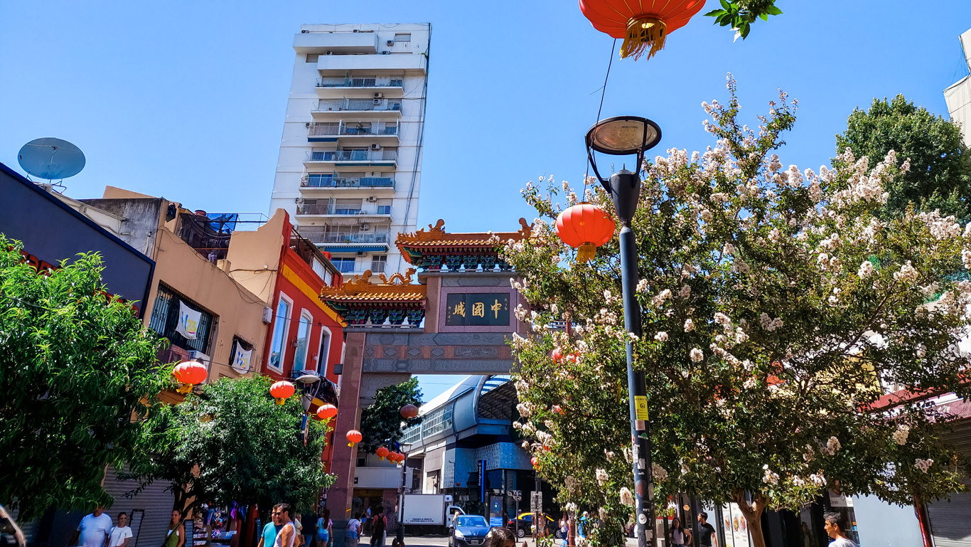 bairro-chino-buenos--aires Entrada del Barrio Chino de Buenos Aires con su portal tradicional, faroles rojos y árboles en flor. Visitantes caminan por las calles estrechas en un día de cielo azul vibrante.