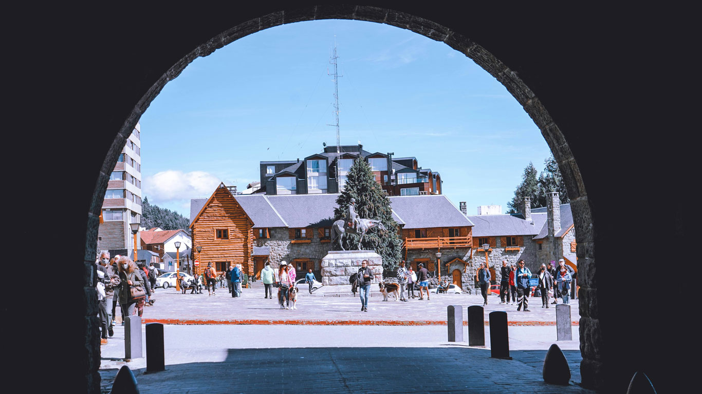 bariloche-en-el-invierno Imagen del Centro Cívico de Bariloche, con su arquitectura de piedra y madera típica y turistas paseando por la plaza, destacando la vida urbana en un destino famoso por la nieve en Bariloche.