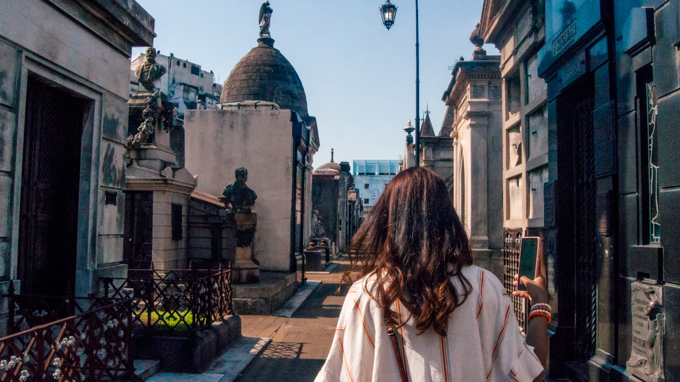 cemiterio-da-recoleta-1 Mujer camina entre las tumbas históricas del Cementerio de la Recoleta, rodeada de esculturas y mausoleos de mármol.