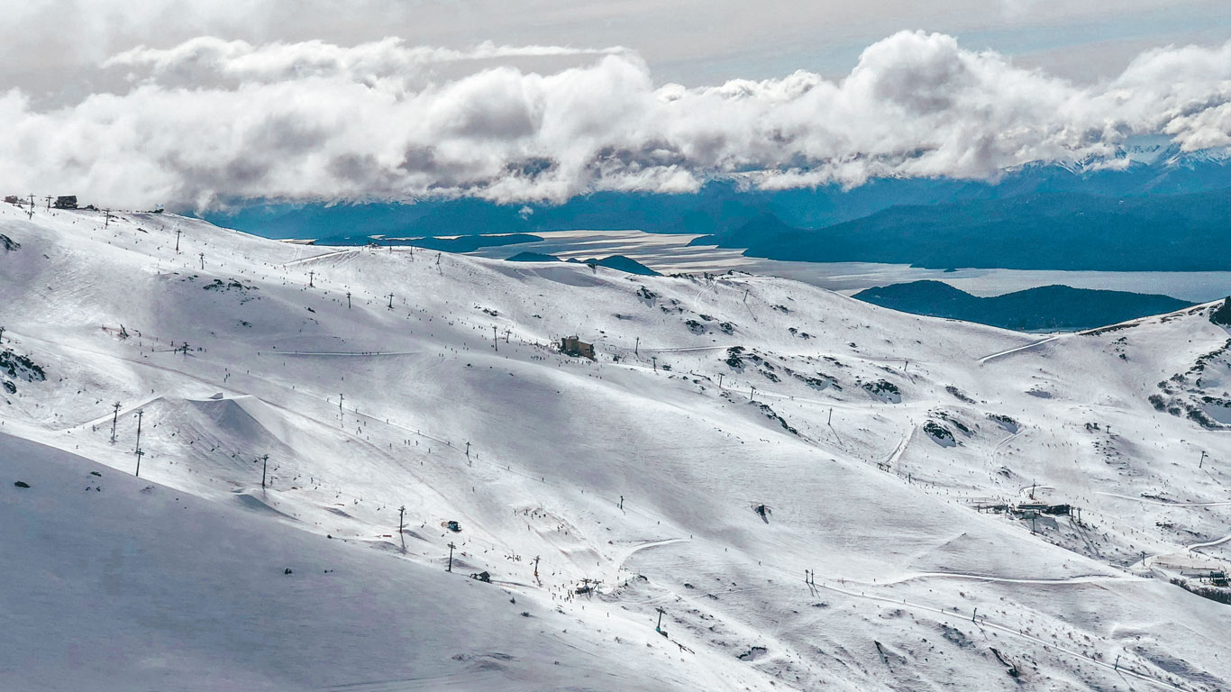 cerro-catedral-1 Extensa vista panorámica del Cerro Catedral cubierto de nieve, con pistas de esquí, telesillas y esquiadores diminutos disfrutando del paisaje típico de nieve en Bariloche.