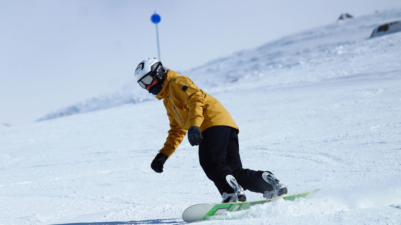 cerro-catedral-bariloche Persona haciendo snowboard en las pistas nevadas del Cerro Catedral, Bariloche, vestida con chaqueta amarilla y casco blanco, deslizándose con estilo sobre la nieve brillante. El cielo despejado y el entorno montañoso refuerzan el ambiente típico de aventura invernal y turismo activo en Bariloche.