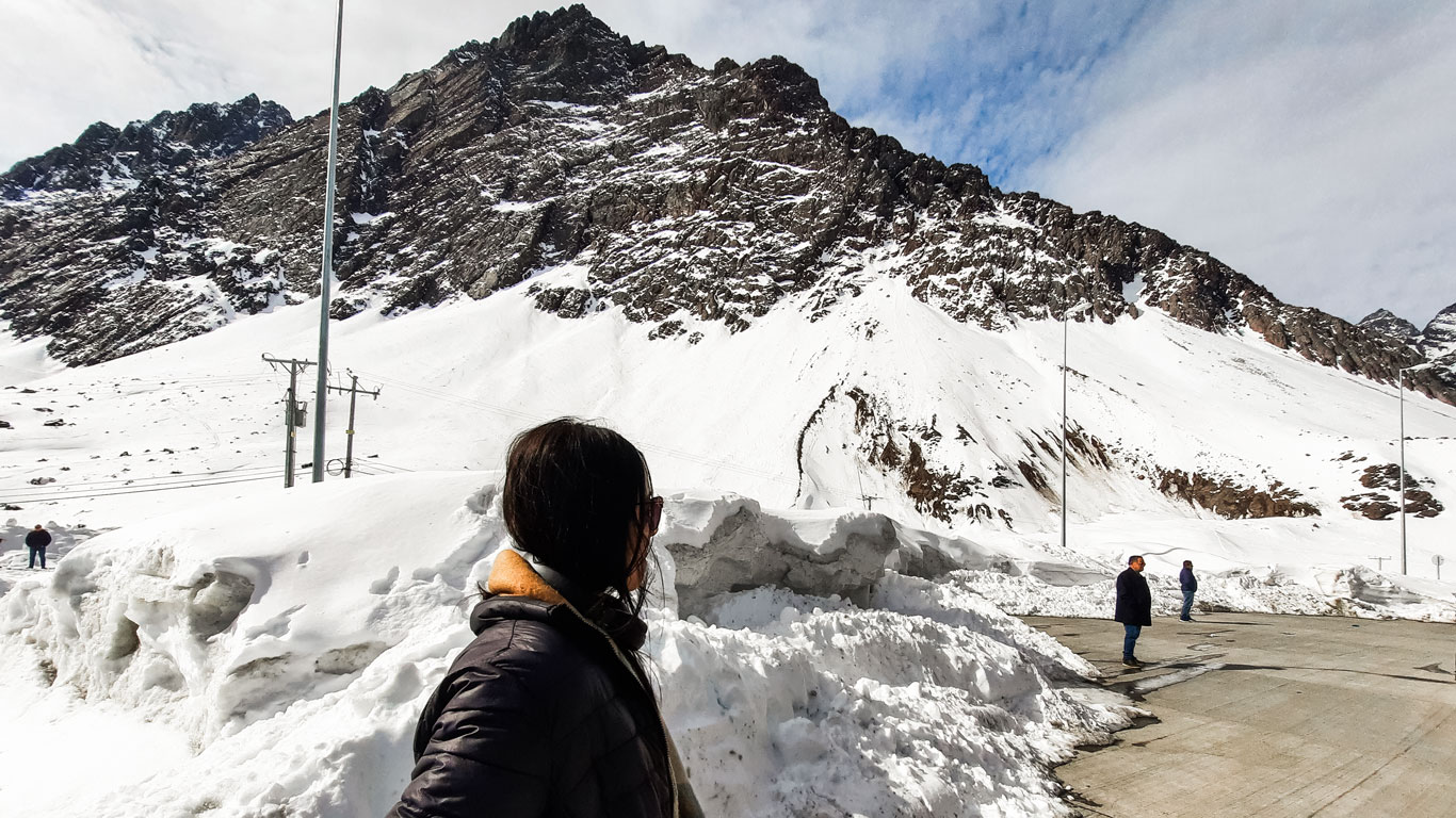 Una persona observa la imponente montaña nevada desde una carretera con nieve acumulada en los bordes. Al fondo, más visitantes disfrutan del paisaje andino bajo un cielo parcialmente nublado.