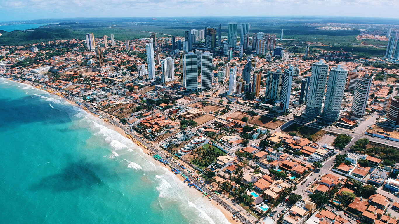 Vista aérea de la ciudad de Natal, Brasil, mostrando la franja costera con edificios modernos, hoteles y sombrillas alineadas en la playa.