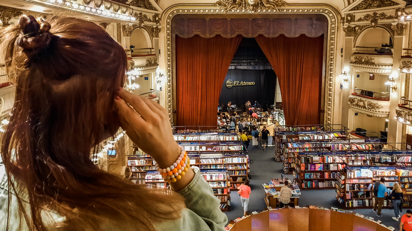 el-ateneo-buenos-aires Mujer observa desde el entrepiso la famosa librería El Ateneo, ubicada en un antiguo teatro con cortinas rojas en el escenario y estanterías de libros repartidas por lo que antes era la platea. El espacio conserva detalles originales de la arquitectura, como adornos dorados y balcones interiores.