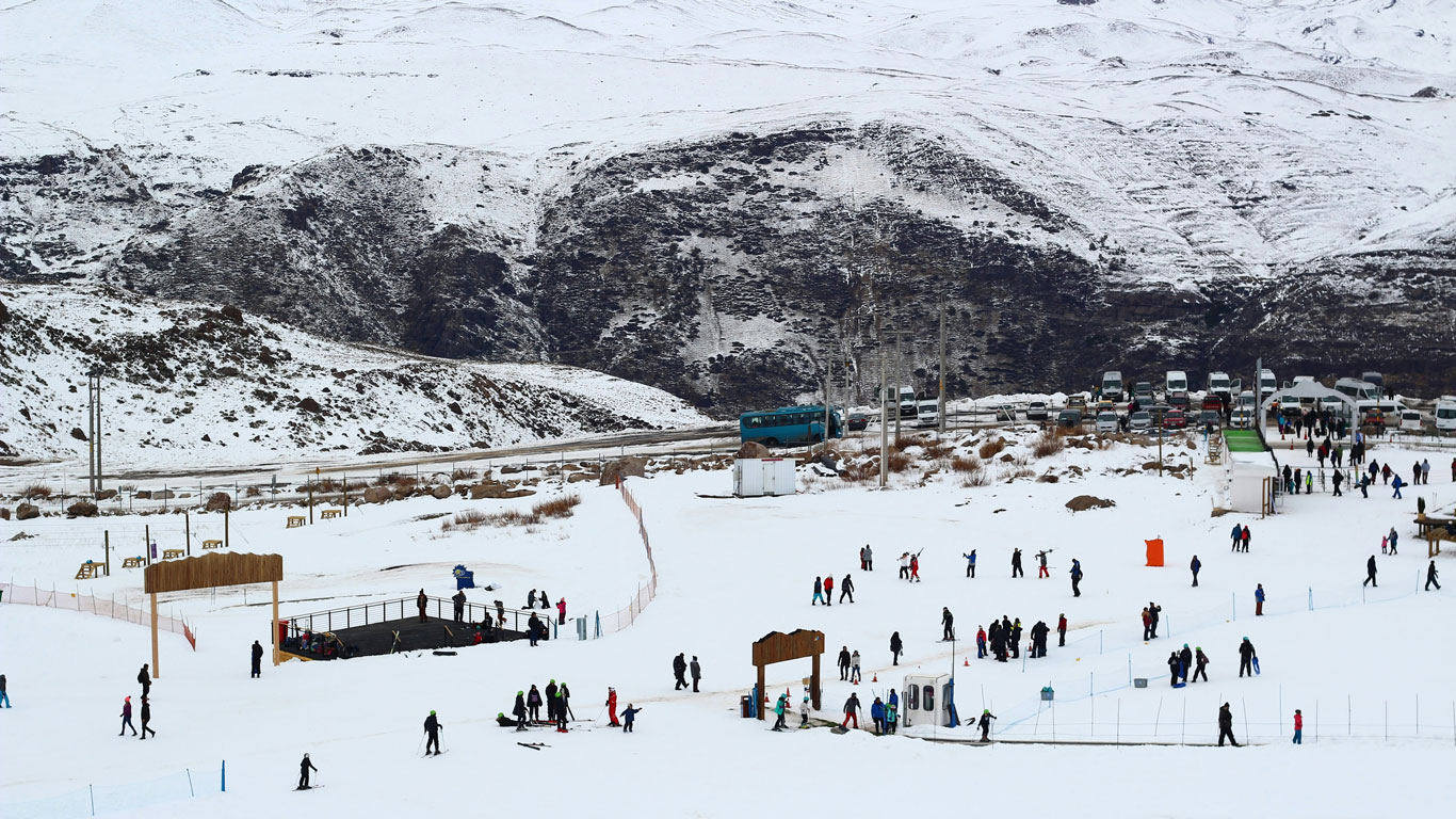 farellones-chile Zona de juegos y deportes de invierno en Farellones, repleta de personas disfrutando de la nieve en Santiago. Al fondo, colinas nevadas, vehículos aparcados y un ambiente familiar típico de la temporada.