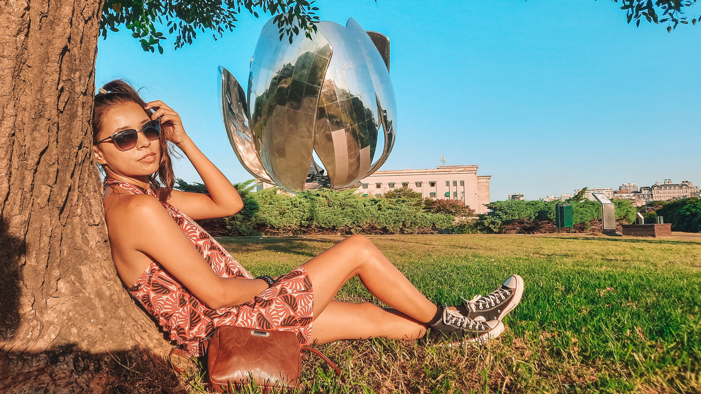 floralis-generica-recoleta Mujer relajada apoyada en un árbol en el parque de la Floralis Genérica, escultura metálica con forma de flor ubicada en Buenos Aires. El día soleado resalta los tonos vibrantes del paisaje.