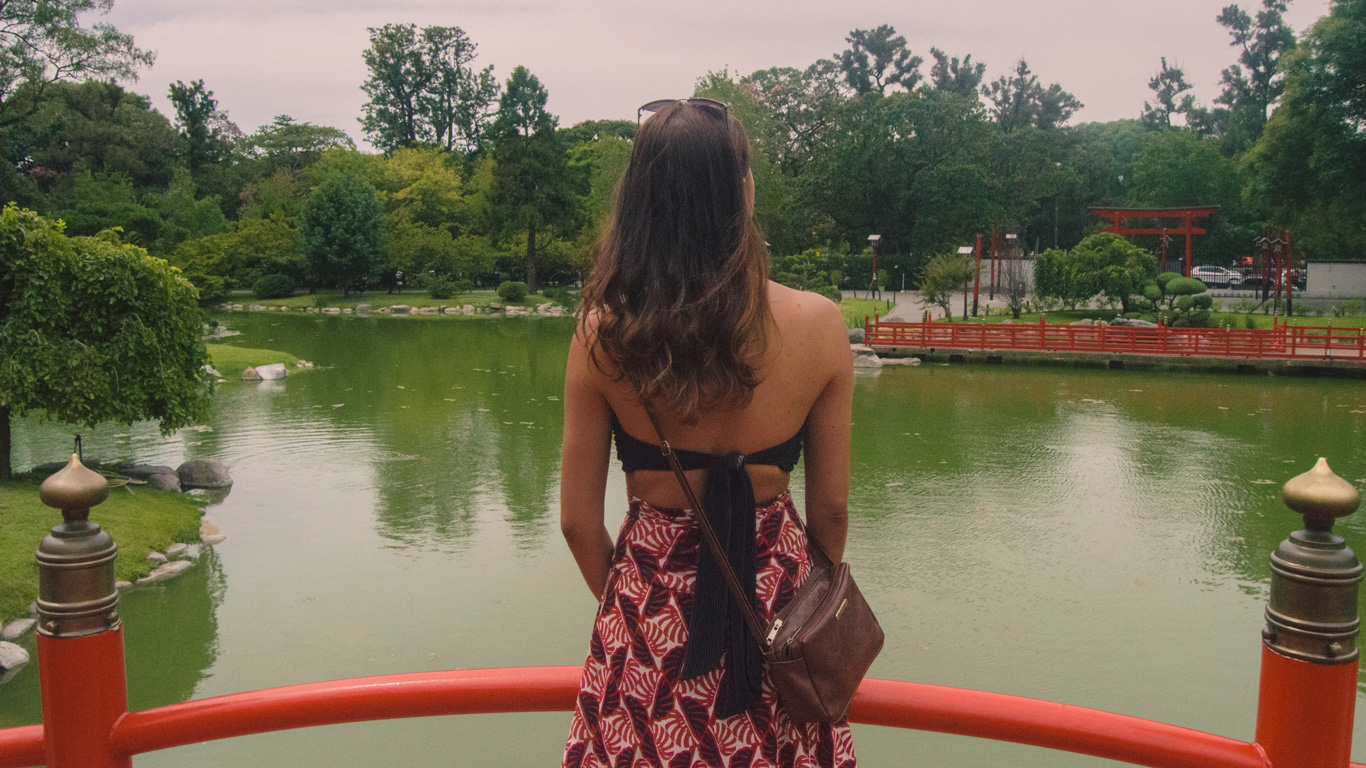 jardin-japones Una mujer de espaldas, con una falda estampada y un top negro, contempla el paisaje tranquilo del Jardín Japonés de Buenos Aires. El lago de reflejos verdosos y las estructuras rojas aportan un toque oriental al entorno.