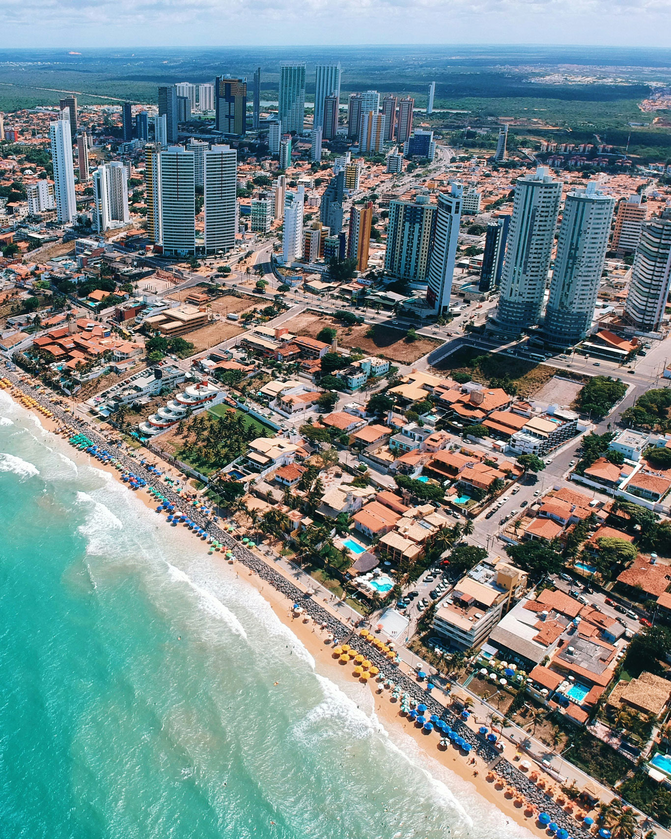 mejor-zona-para-hospedarse-en-natal Otra vista aérea de Natal mostrando una gran concentración de rascacielos frente al mar, con muchas sombrillas a lo largo de la playa. Representa visualmente la mejor zona donde alojarse en Natal si se desea acceso directo a la playa y servicios turísticos.