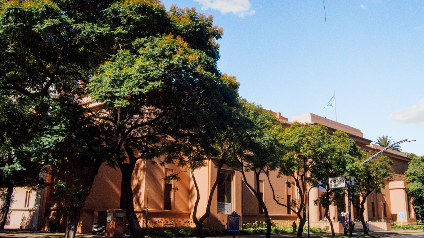 museu-nacional-de-belas-artes Fachada del Museo Nacional de Bellas Artes, rodeada de árboles y vegetación frondosa. El edificio en tono terracota luce la bandera de Argentina y destaca bajo un cielo azul con pocas nubes.