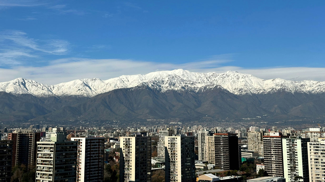 nieve-in-santiago-cordillera-de-los-andes Vista panorámica de Santiago con edificios modernos en primer plano y la Cordillera de los Andes nevada al fondo, bajo un cielo despejado. Una imagen icónica que representa la belleza de la nieve en Santiago durante el invierno.