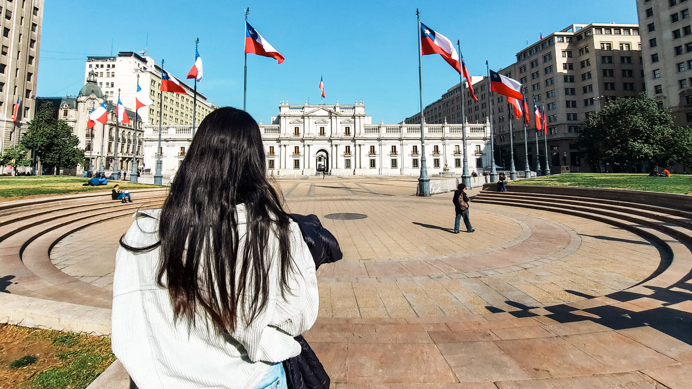 palacio-de-la-moneda-santiago Turista observa el Palacio de La Moneda en Santiago, rodeado de banderas chilenas y un cielo soleado.