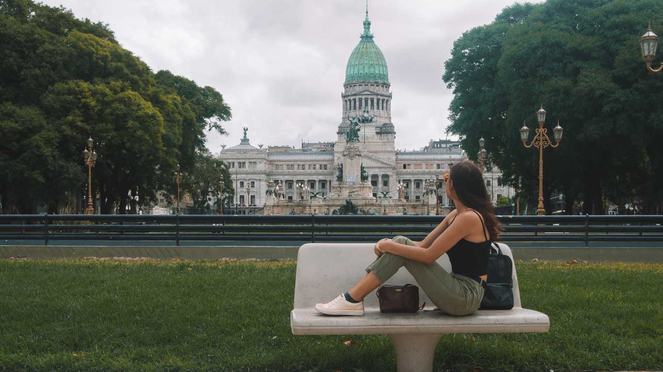 plaza-del-congresso Mujer sentada en un banco de la plaza observa el edificio del Congreso Nacional, con su cúpula verde y arquitectura imponente. Árboles rodean el entorno urbano e institucional.