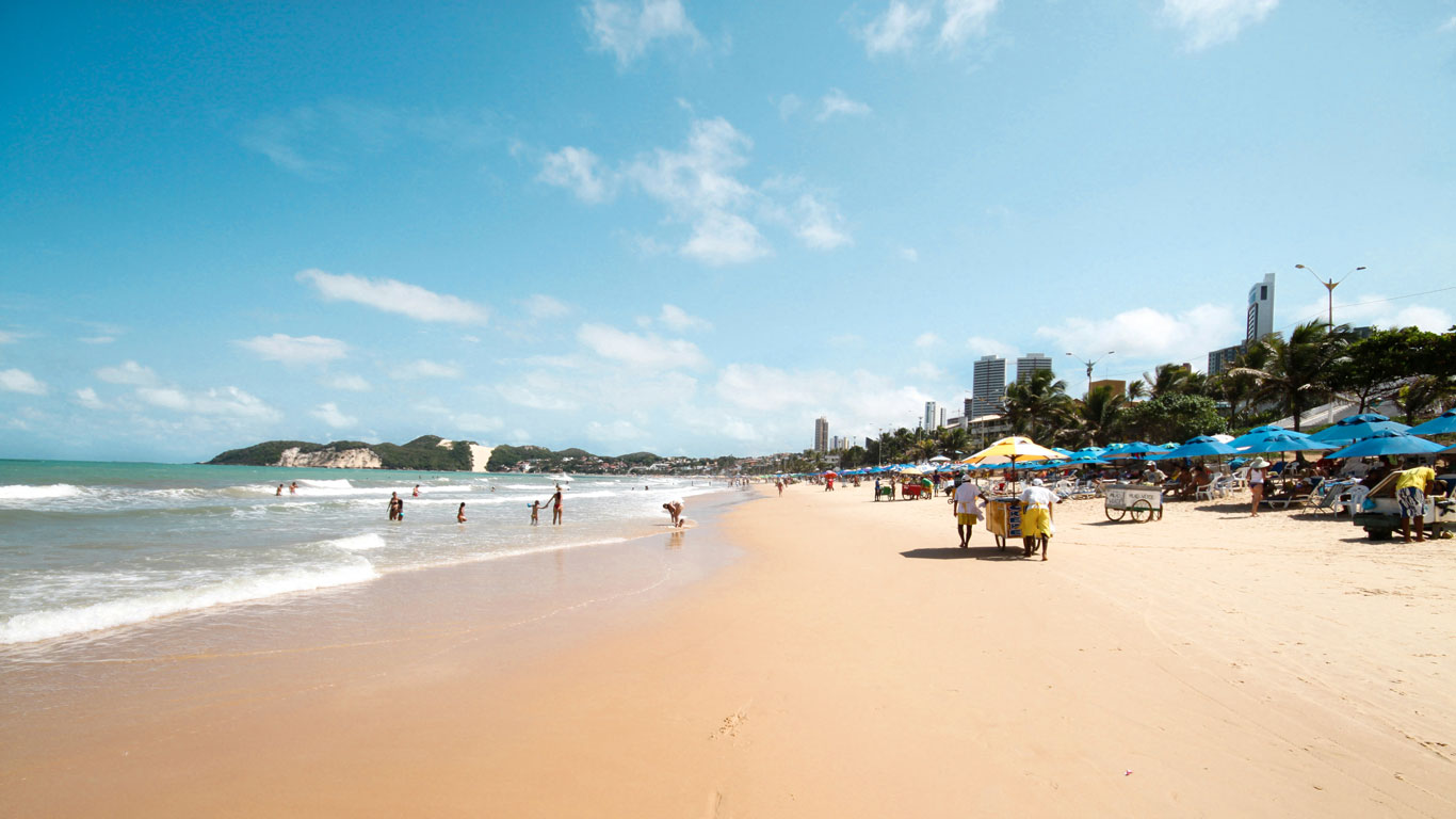 ponta-negra-natal Playa de Ponta Negra en Natal, con sombrillas de colores, turistas disfrutando del mar y edificios al fondo. Esta zona es una de las respuestas favoritas a la pregunta de donde alojarse en Natal, ideal para quienes buscan ambiente y comodidad.