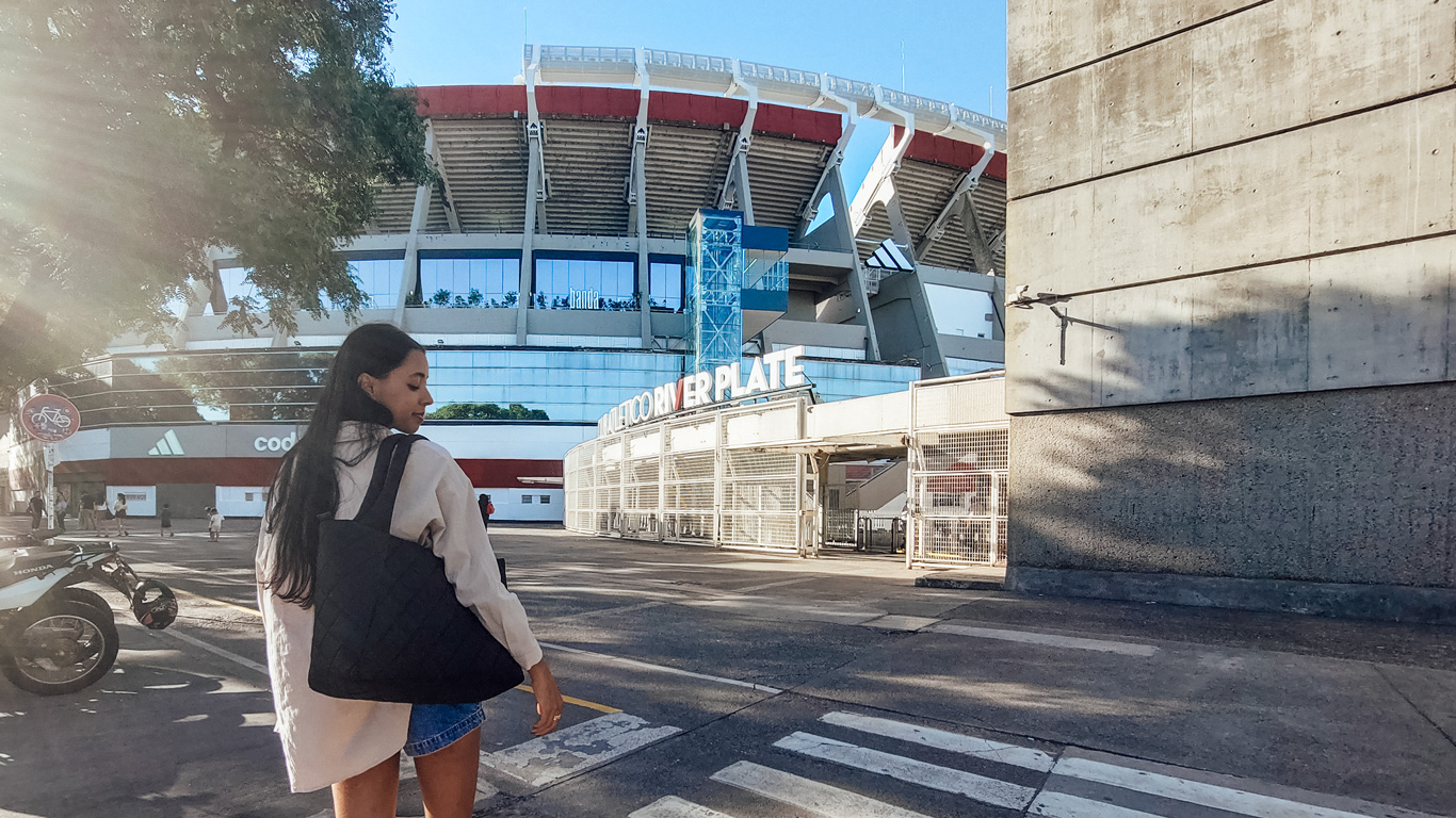river-plate Mujer con ropa informal camina frente al estadio Monumental de Núñez, casa del River Plate. La arquitectura moderna y el clima soleado completan el paisaje urbano del lugar.