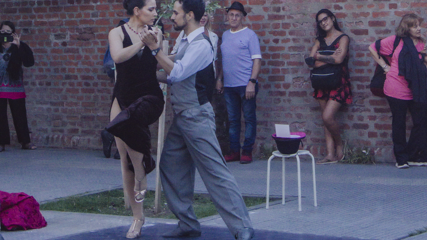 tango-en-buenos-aires Pareja se presenta con el clásico look de tango en las calles de Buenos Aires; frente al Cementerio de la Recoleta, ella lleva un vestido negro con abertura y tacones, mientras él viste camisa y chaleco gris. Al fondo, varias personas observan la actuación intensa y apasionada.