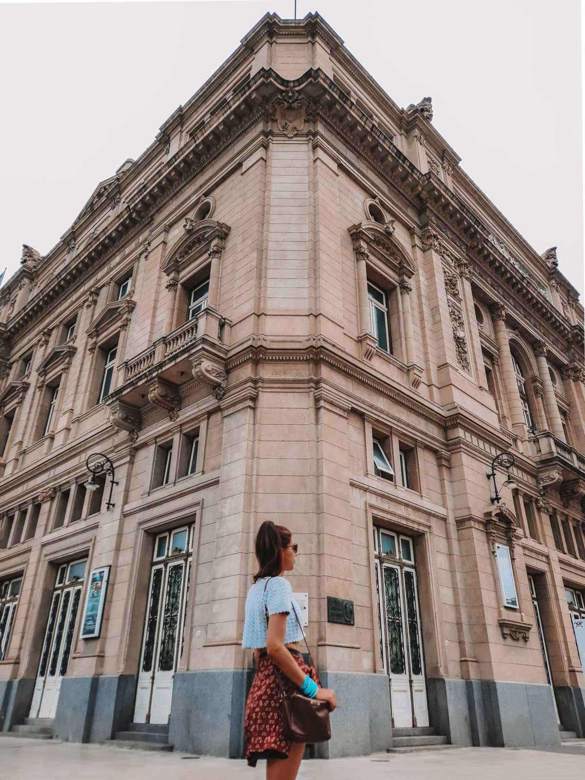 teatro-colon-1 La imagen muestra a una mujer parada frente al Teatro Colón, en Buenos Aires, contemplando la imponente fachada del edificio. El teatro, con su arquitectura clásica y detalles ornamentados, destaca por sus ventanas y molduras elegantes. La mujer lleva una blusa blanca y una falda estampada, y sostiene un bolso mientras admira la majestuosidad del edificio histórico.