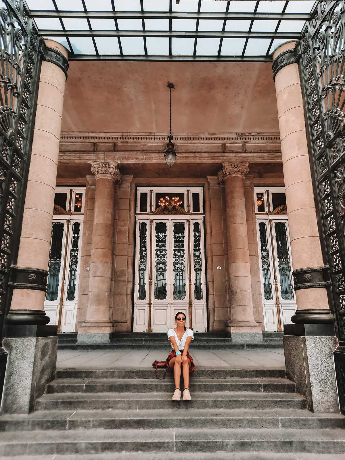 teatro-colon La imagen también muestra la entrada del Teatro Colón en Buenos Aires, con una mujer sentada en los escalones frontales. La estructura está adornada con columnas clásicas y rejas de hierro forjado, con altas puertas al fondo. El techo de cristal y las lámparas de época completan la imponencia arquitectónica del lugar. La mujer sonríe y va vestida de manera informal, aportando un aire relajado a la escena.