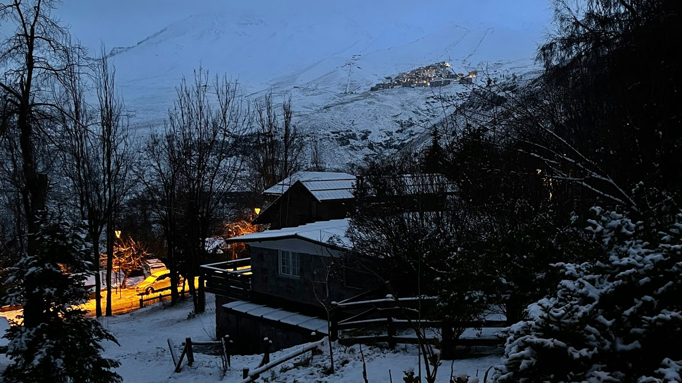 valle-nevado-chile Una escena nocturna en una aldea de montaña nevada, con cabañas de madera iluminadas tenuemente y farolas anaranjadas. Al fondo, una ladera montañosa cubierta de nieve y un poblado en lo alto, brillando con luces cálidas.