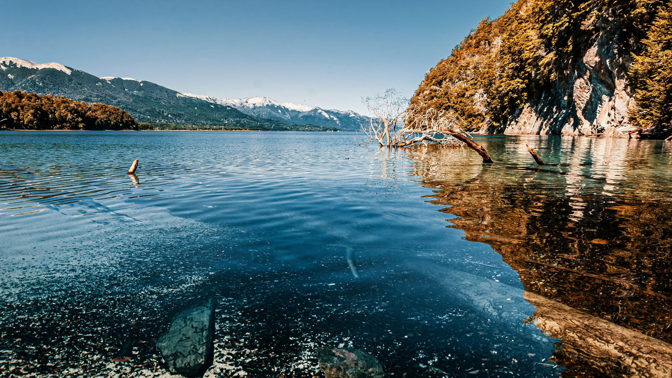 villa-la-angostura Aguas cristalinas y reflejos de árboles y montañas en un lago patagónico, bajo un cielo despejado. Una postal que también forma parte del recorrido hacia la nieve en Bariloche durante el invierno.