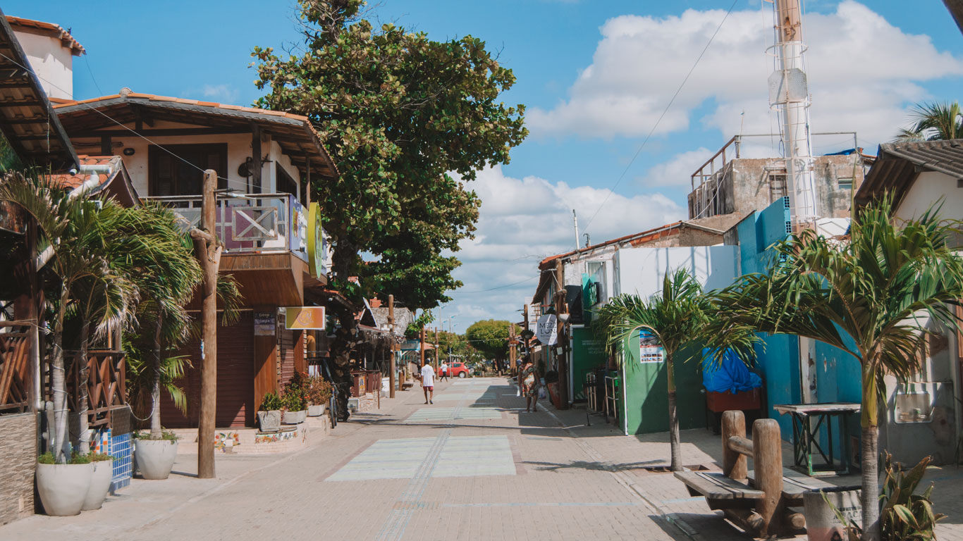 Calle con encanto y ambiente tranquilo, con tiendas y posadas de estilo rústico, flanqueada por palmeras y árboles bajo un cielo azul con nubes, en el centro, el mejor lugar para alojarse en Canoa Quebrada. Al fondo, algunas personas pasean en un ambiente relajado.
