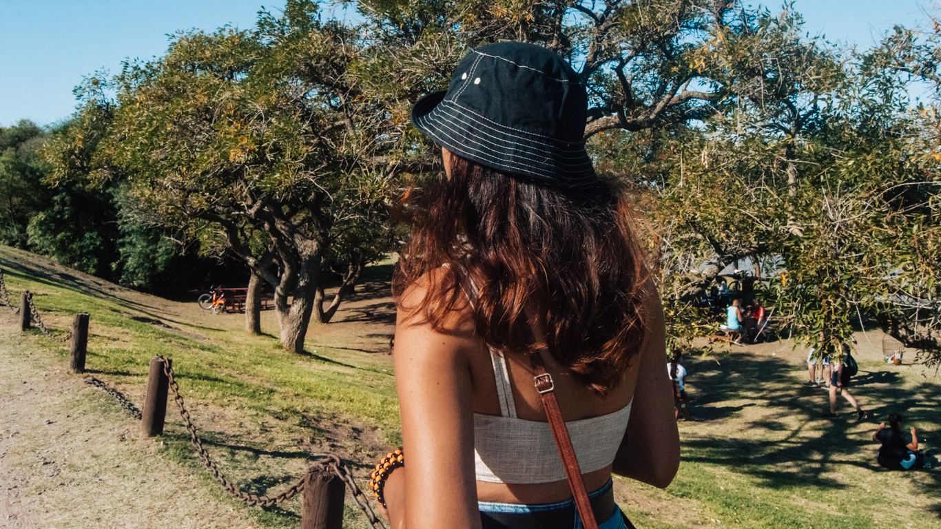 costanera-sur-buenos-aires Una mujer con sombrero oscuro pasea por la Reserva Ecológica Costanera Sur, un parque arbolado en un día soleado, rodeada de personas descansando sobre el césped.