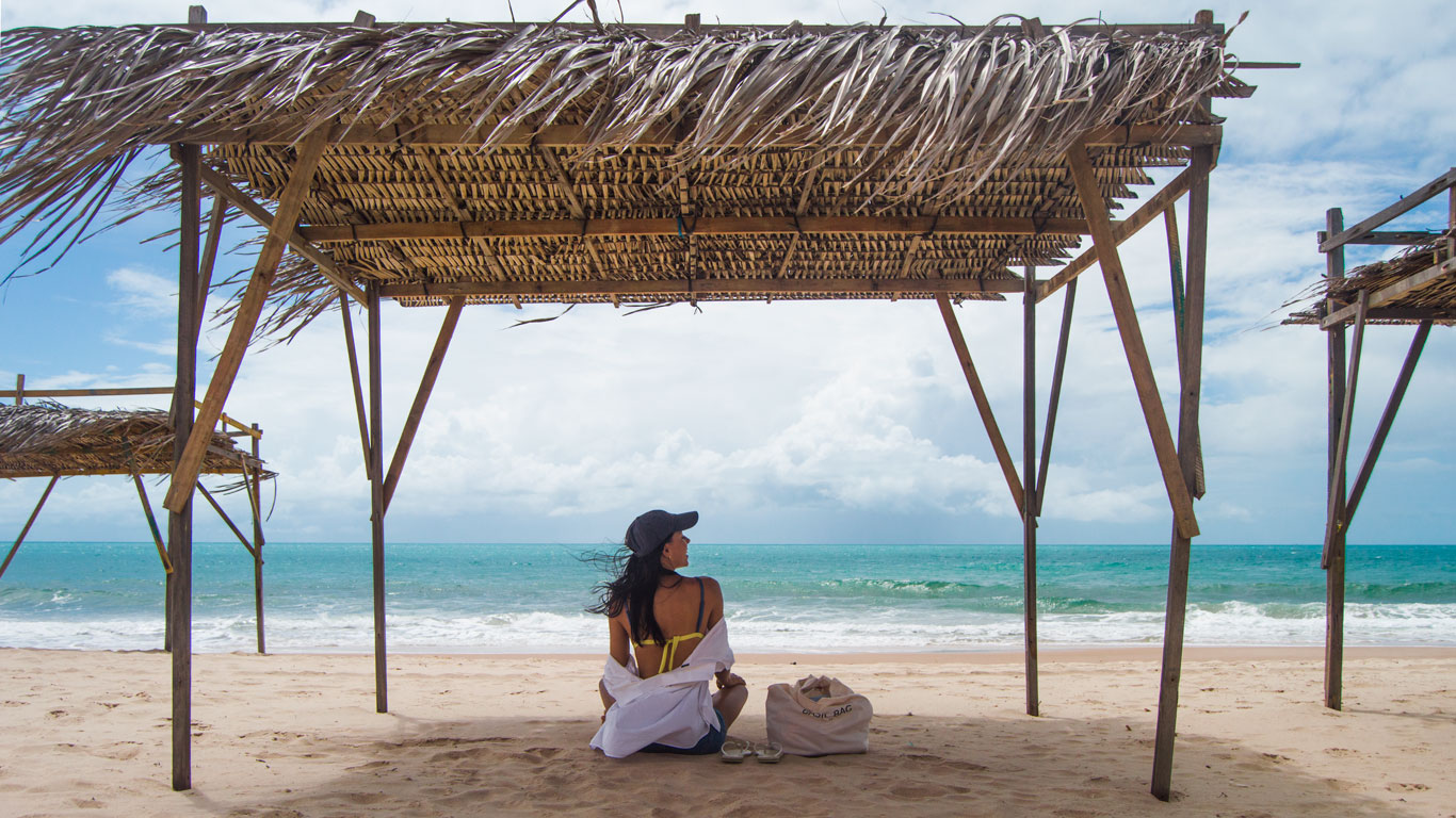Mujer sentada en la arena bajo un chiringuito rústico de madera con techo de paja, mirando el mar azul de Canoa Quebrada. A su lado, una bolsa de playa y unas chanclas, en un ambiente de tranquilidad junto al mar.