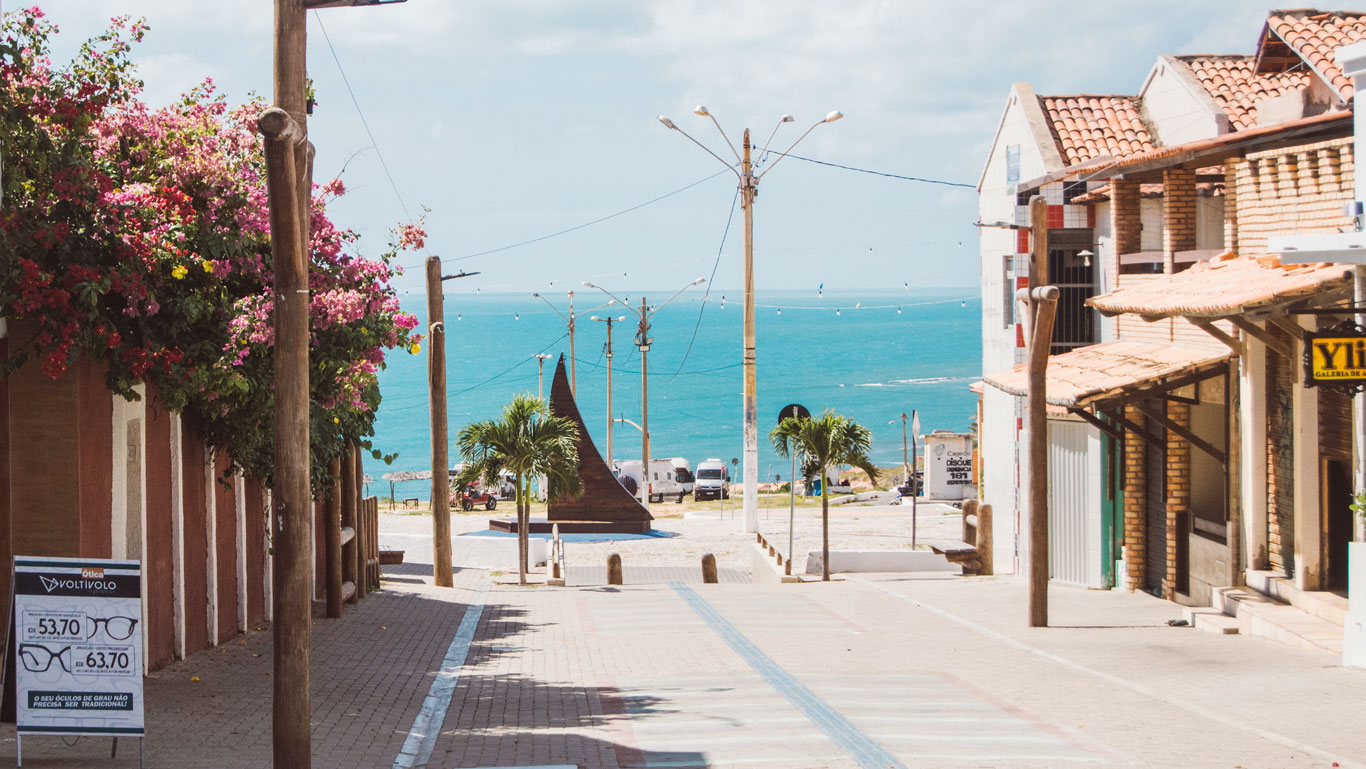 Vista de la calle Broadway en el centro de Canoa Quebrada, que desciende hacia el mar azul, con postes, palmeras y algunas construcciones de ladrillo y carteles comerciales. Al fondo se ve una escultura con forma de vela náutica.