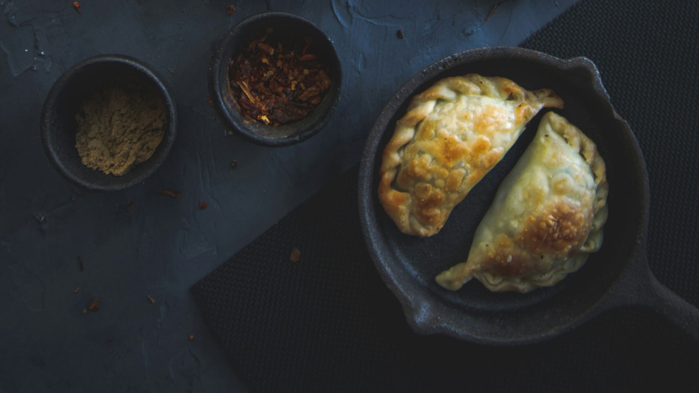 Empanadas argentinas doradas servidas en una sartén de hierro junto a cuencos con especias secas, sobre una superficie oscura.