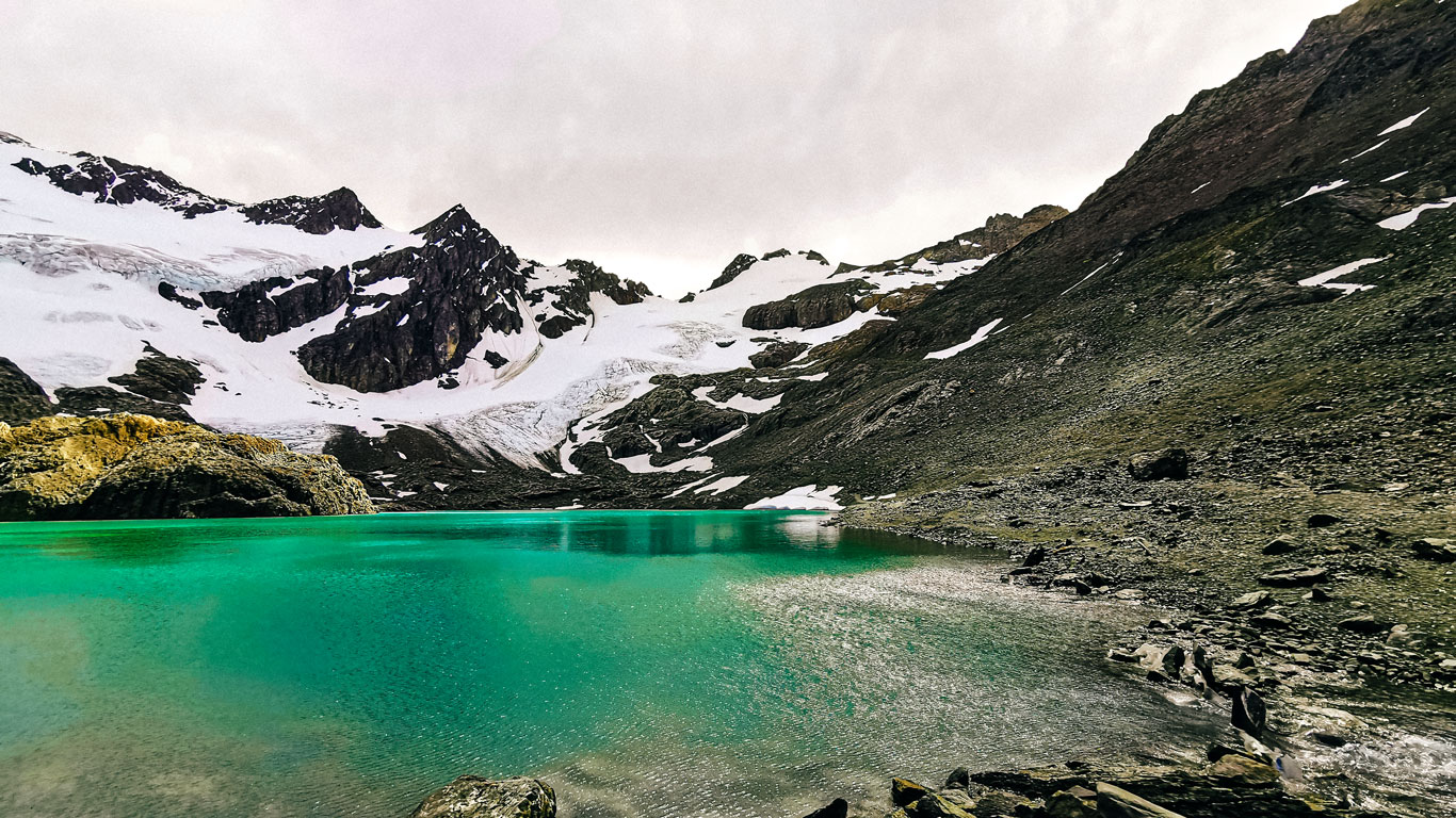 Lago de aguas verde esmeralda reflejando la luz bajo un cielo nublado, rodeado de laderas rocosas y el glaciar Vinciguerra parcialmente cubierto de nieve en los alrededores de Ushuaia.