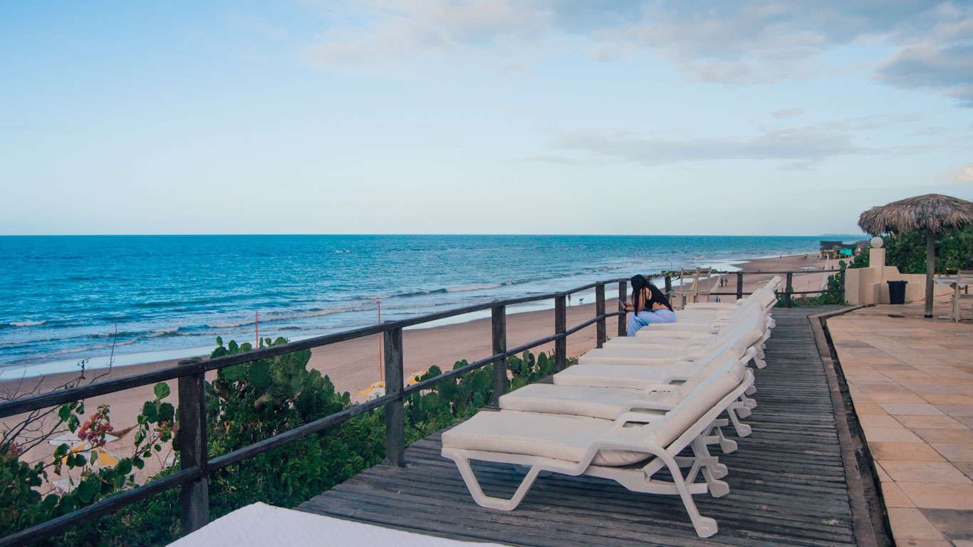 Terraza de madera con tumbonas blancas frente a la playa, en la Pousada Vila Canoa, donde una mujer está sentada mirando el mar azul bajo un cielo despejado.