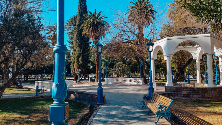 Plaza Independencia en Mendoza en octubre, arbolada con bancos de madera, farolas azules y un quiosco blanco, bajo un cielo despejado y azul.