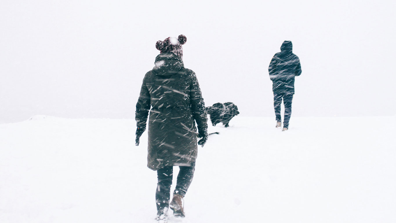 Dos personas y un perro caminando sobre la nieve durante una nevada, con ropa de abrigo oscura que contrasta con el blanco del entorno.